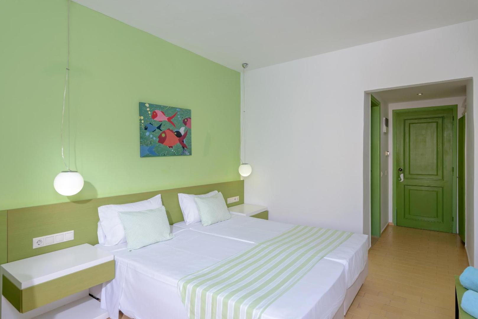 Zeus Palm Beach Hotel gaišs divvietīgs numurs ar zaļu akcenta sienu, modernām piekaramām lampām un baltu gultas veļu Stalidā, Krētā.