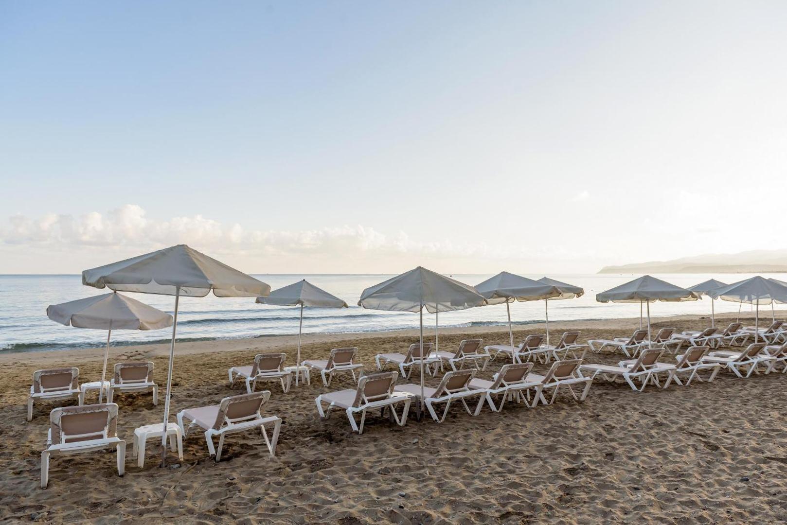 Zeus Palm Beach Hotel privātā smilšu pludmale Krētā ar gaišiem saulessargiem un sauļošanās krēsliem pie mierīgas jūras rīta gaismā