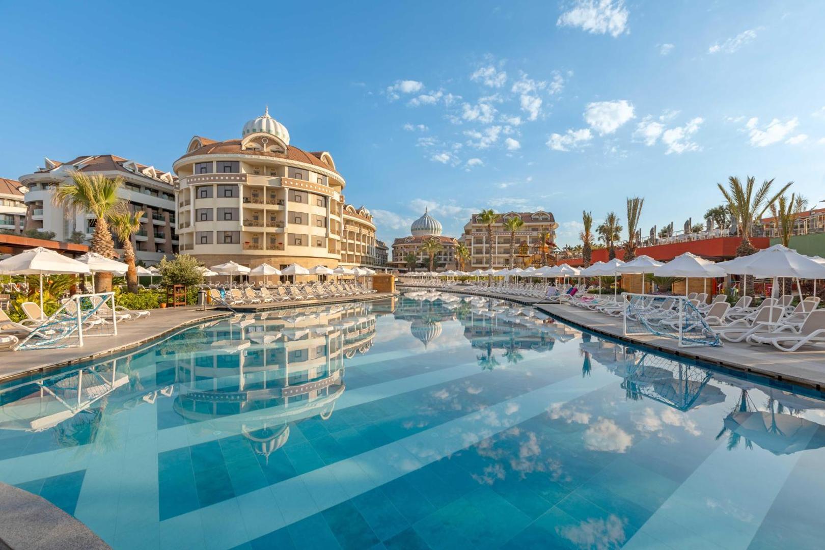 Kirman Hotels Belazur Resort & Spa lielais āra baseins ar saulessargiem un viesnīcas korpusiem fonā Belekā, Turcijā saulainā dienā
