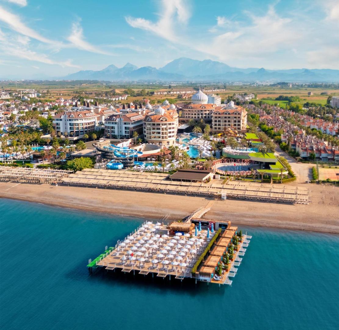 Kirman Belazur Resort & Spa panorāmas skats no putna lidojuma Belekā — viesnīcas komplekss, baseini, smilšu pludmale un plaša koka piestātne ar saulessargiem