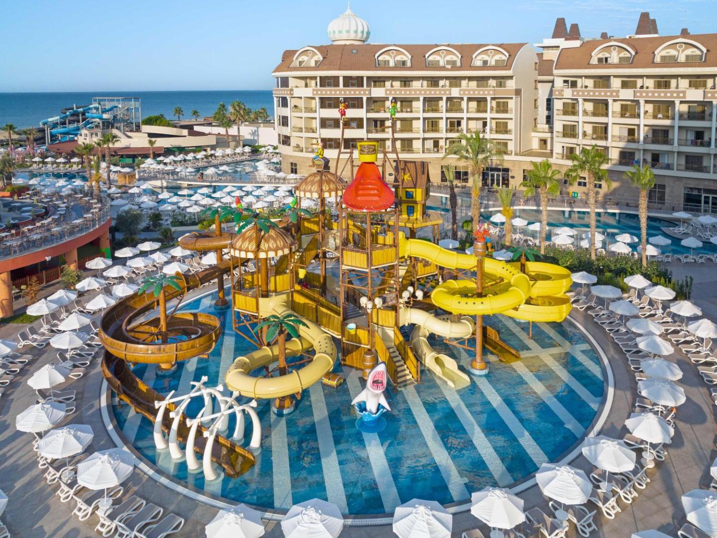 Kirman Hotels Belazur Resort & Spa bērnu akvaparks ar krāsainiem slidkalniņiem un baseinu Belekā, Turcijā — viesnīcas ēka un sauļošanās krēsli
