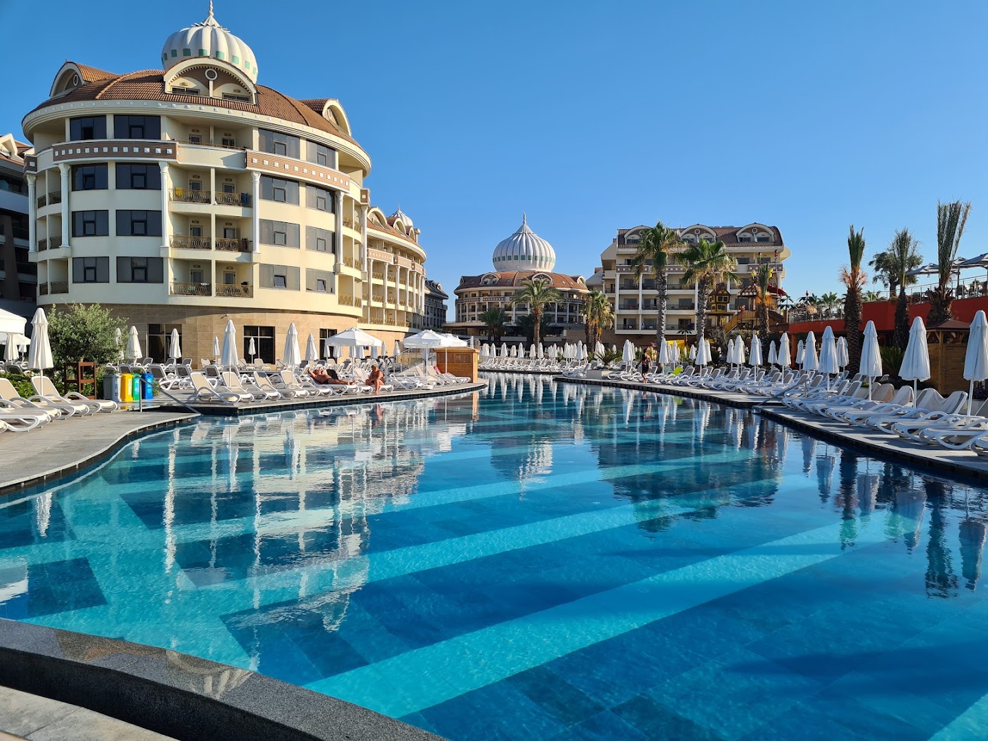 Skats uz Kirman Hotels Belazur Resort & Spa baseinu un viesnīcas arhitektūru ar kupoliem zem skaidrām debesīm Belekā
