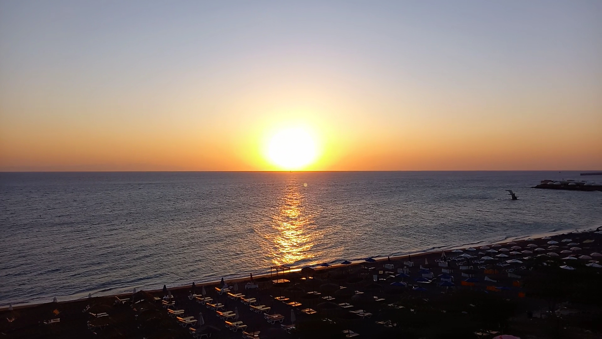 Saulriets virs Vidusjūras pie Mediterranean Hotel Rodā — skats uz pludmali ar saulessargiem vakara gaismā