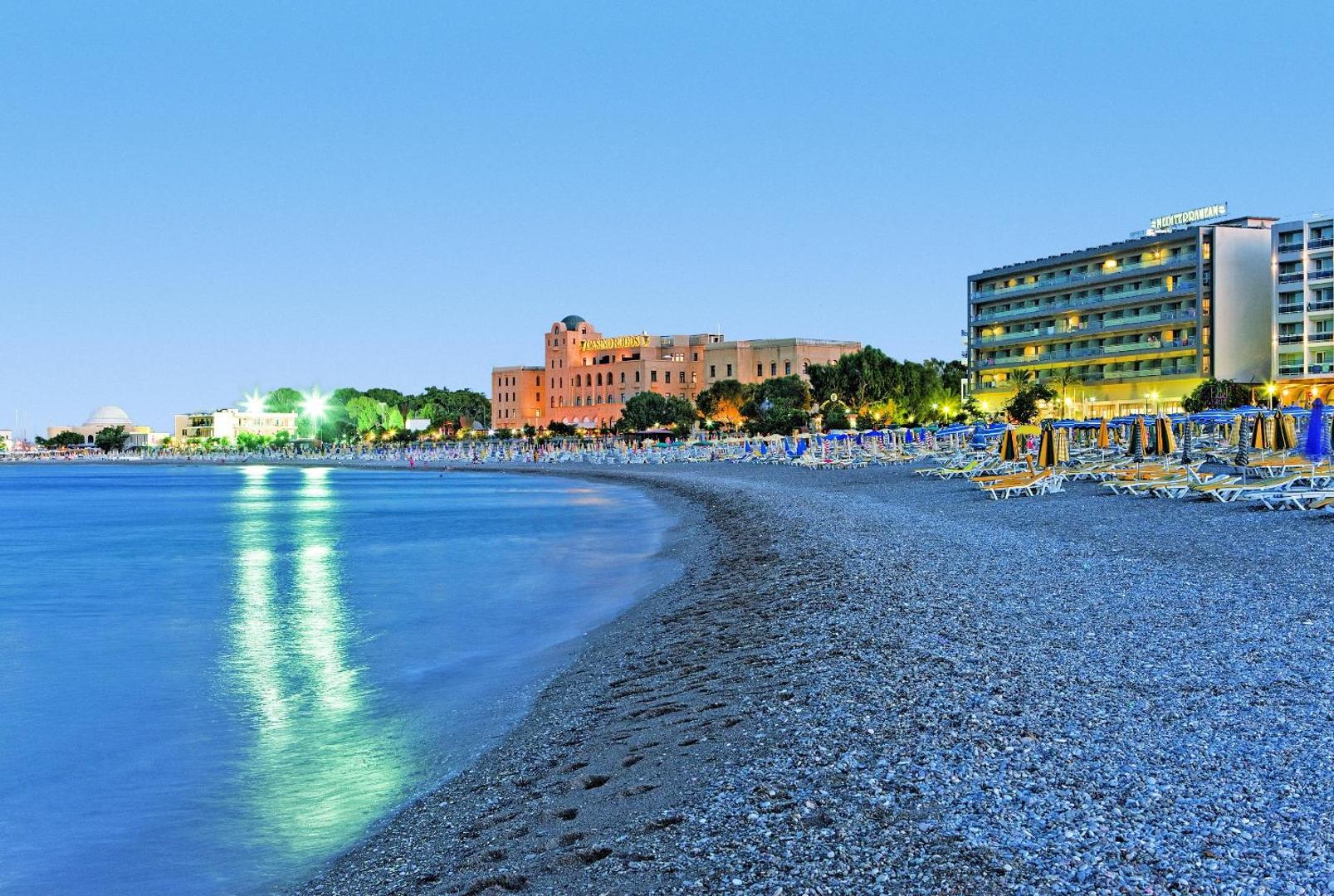 Mediterranean Hotel skats uz oļu pludmali un pilsētas panorāmu vakarā Rodas salā, Grieķijā — izgaismota krasta līnija ar atpūtas krēsliem