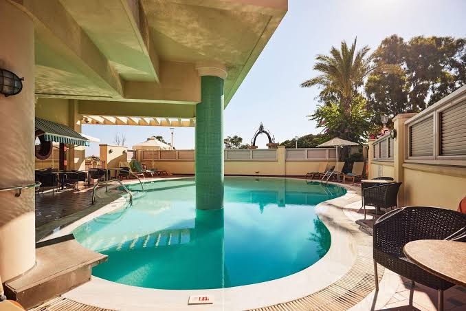 Mediterranean Hotel āra baseins ar tirkīzzilu ūdeni, atpūtas krēsliem un saulessargiem Rodas salas piekrastē