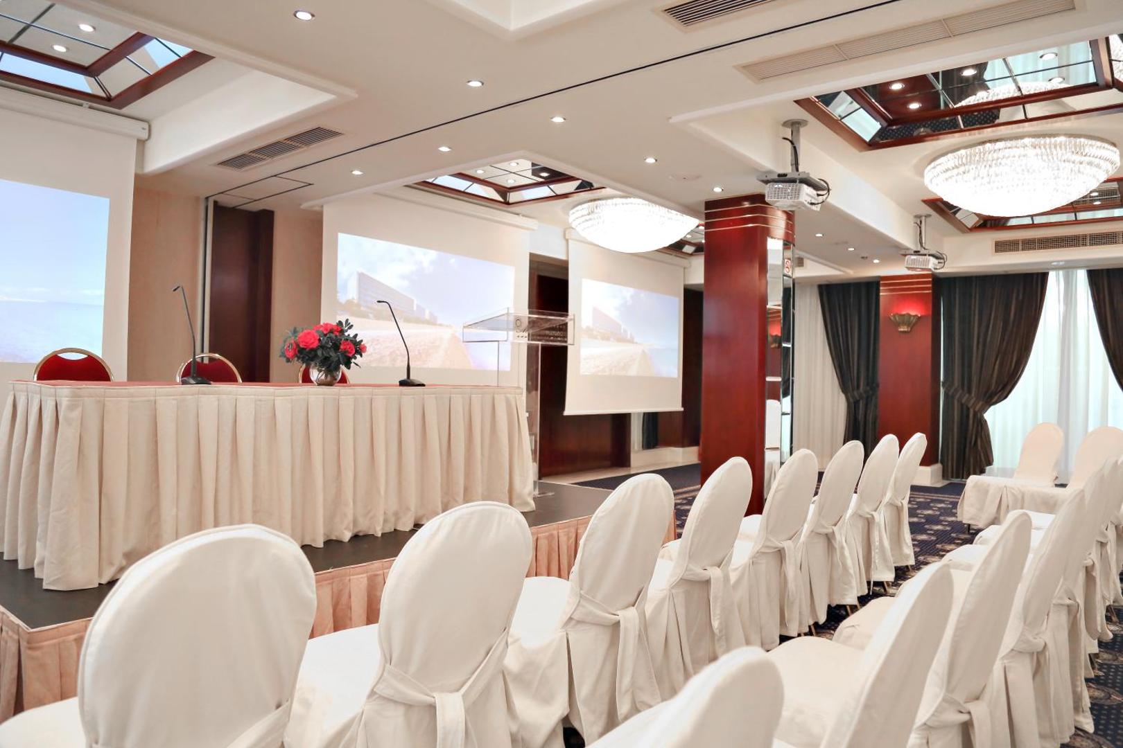 Mediterranean Hotel konferenču zāle Rodas salā ar baltiem krēslu pārvalkiem, projektoru ekrāniem un svinīgu galdu prezentācijām