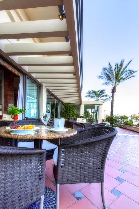 Mediterranean Hotel āra terase ar pītiem krēsliem un galdiņiem — skats uz palmām un saulaino debess pusi Rodas kūrortā