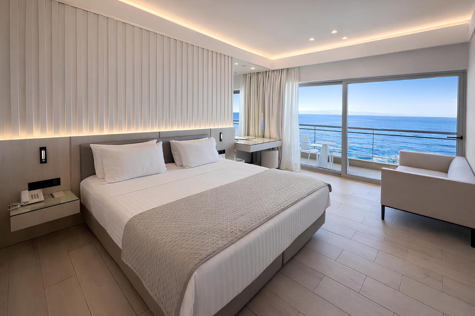 Moderns Mediterranean Hotel divvietīgs numurs ar gaišu interjeru un plašu logu, no kura paveras tiešs skats uz Vidusjūru