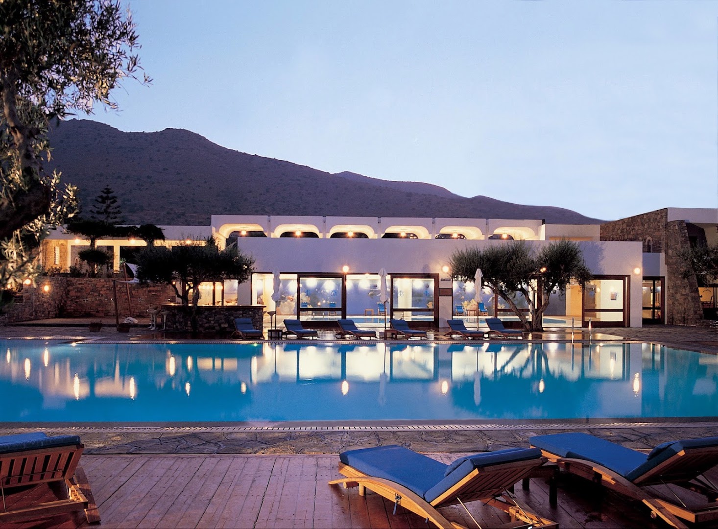 Elounda Bay Palace Hotel izgaismotais āra baseins vakarā ar atpūtas krēsliem un skatu uz kalnu siluetu Krētā