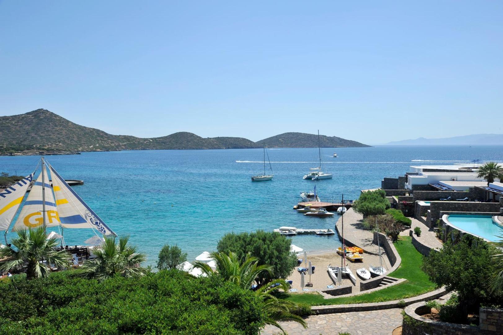 Elounda Bay Palace Hotel panorāmas skats uz Mirabello līci Krētā — viesnīcas teritorija, baseins un jahtas zilajos piekrastes ūdeņos