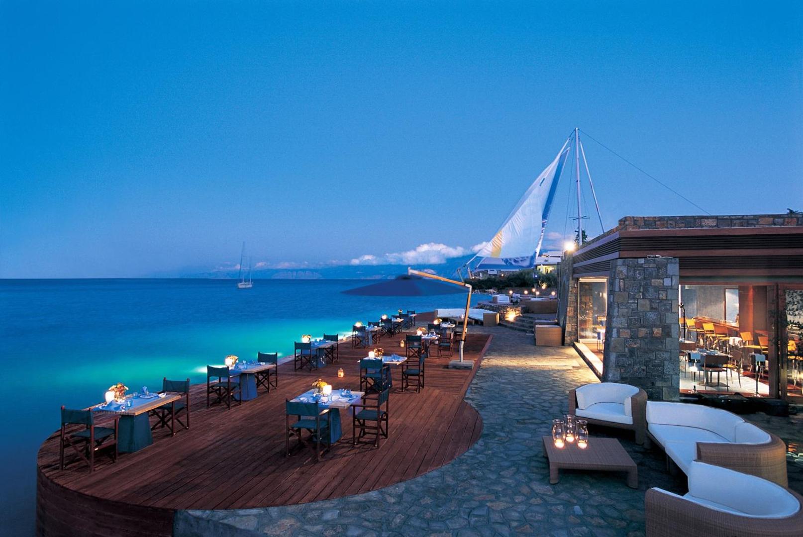 Elounda Bay Palace Hotel restorāna terase vakarā pie jūras Elundā, Krētā — koka klājs ar galdiņiem un buru dekorācijām