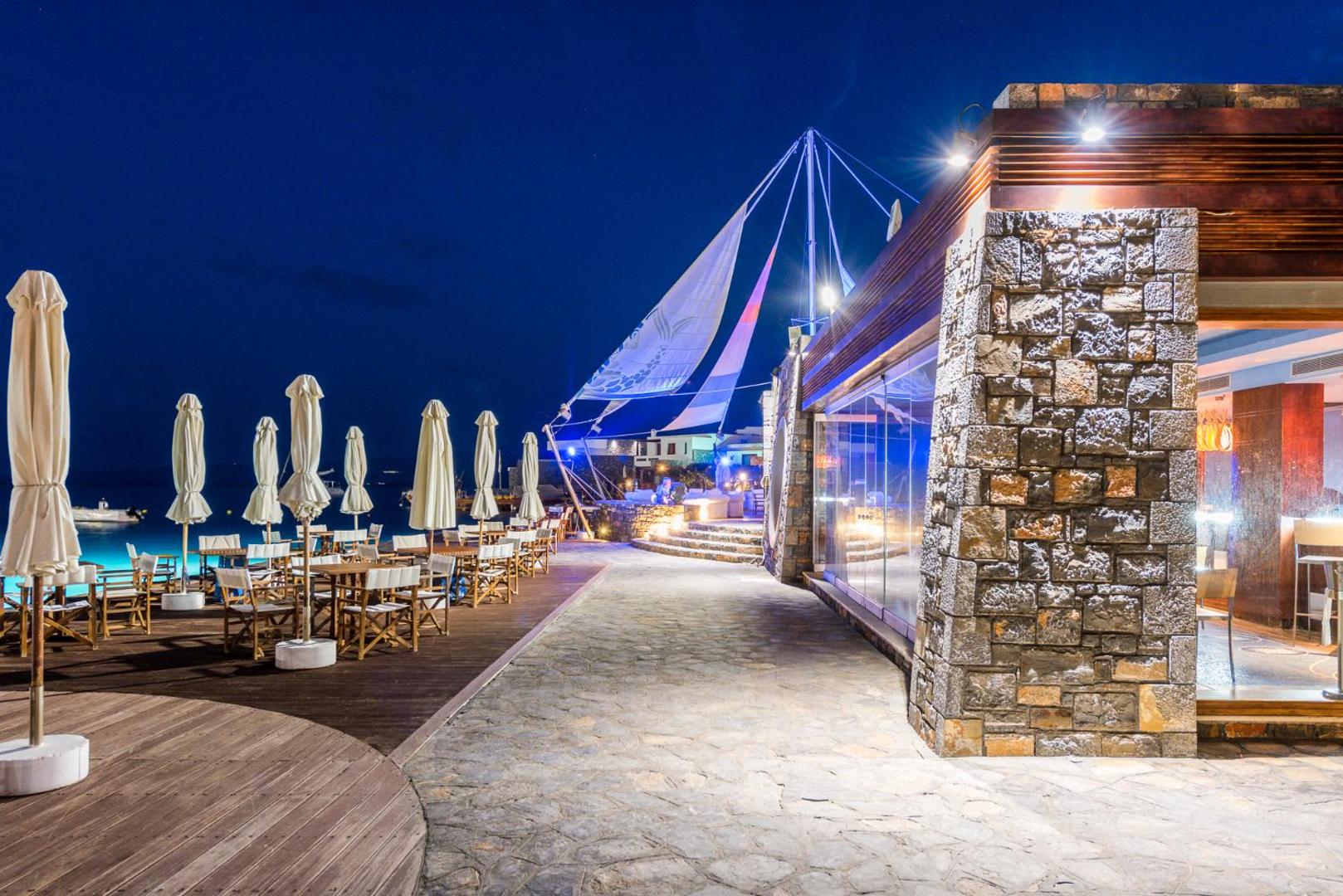 Elounda Bay Palace Hotel restorāna āra terase vakarā ar galdiņiem, saulessargiem un izgaismotu akmens fasādi Krētas piekrastē