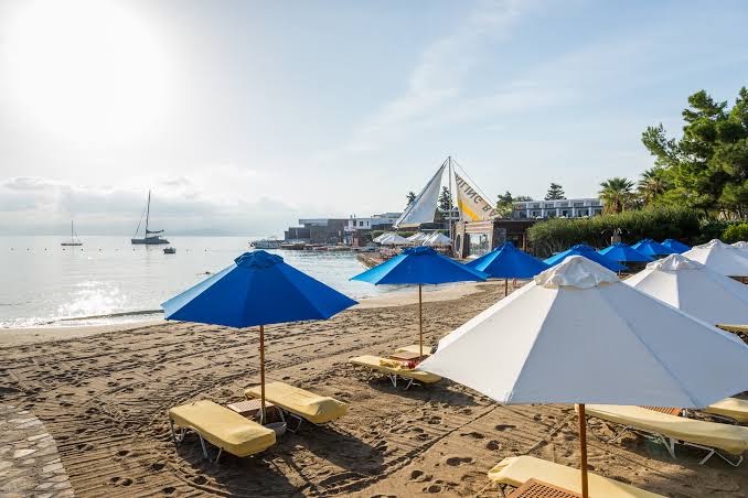 Elounda Bay Palace Hotel labiekārtota pludmale ar saulessargiem un sauļošanās krēsliem smiltīs, fonā redzamas jahtas līcī