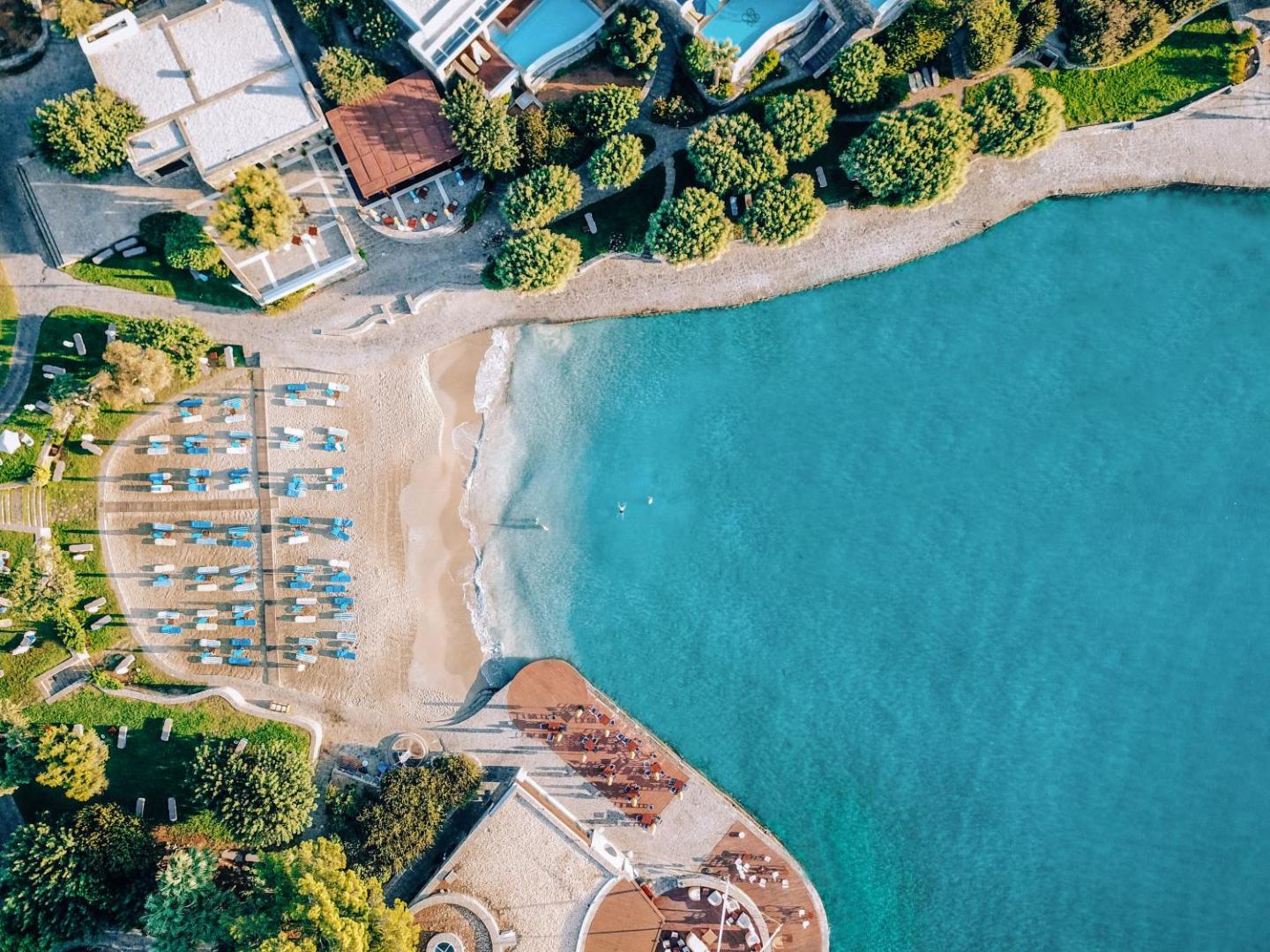 Elounda Bay Palace Hotel skats no putna lidojuma uz smilšu pludmali un tirkīzzilo līci Krētā, Grieķijā ar sauļošanās krēsliem.