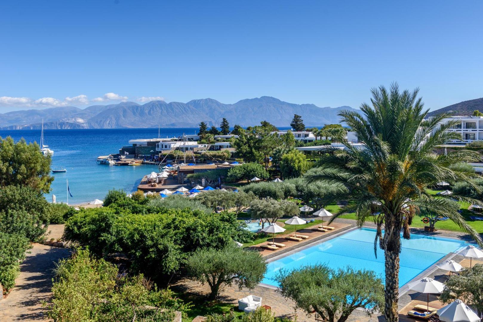 Elounda Bay Palace Hotel teritorijas panorāma ar galveno baseinu, palmām un skatu uz kalniem un jūru Krētas ziemeļos