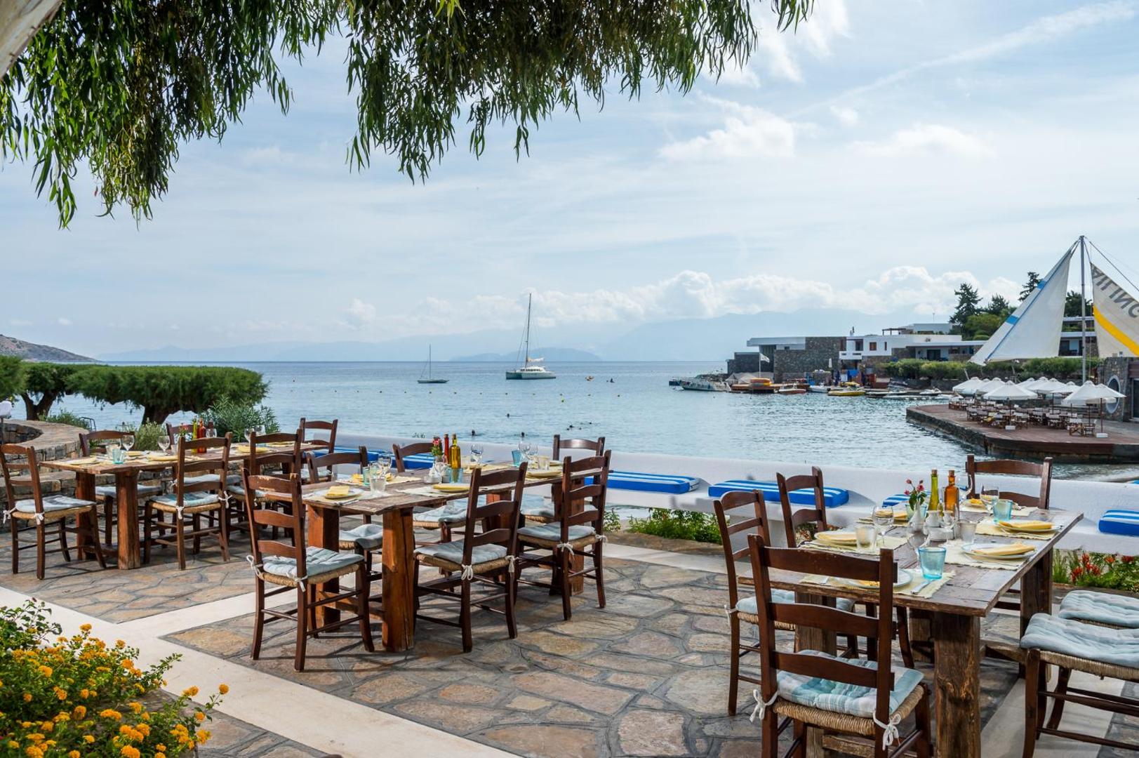 Elounda Bay Palace Hotel brīvdabas restorāns piekrastē ar koka galdiem un skatu uz jahtām līcī