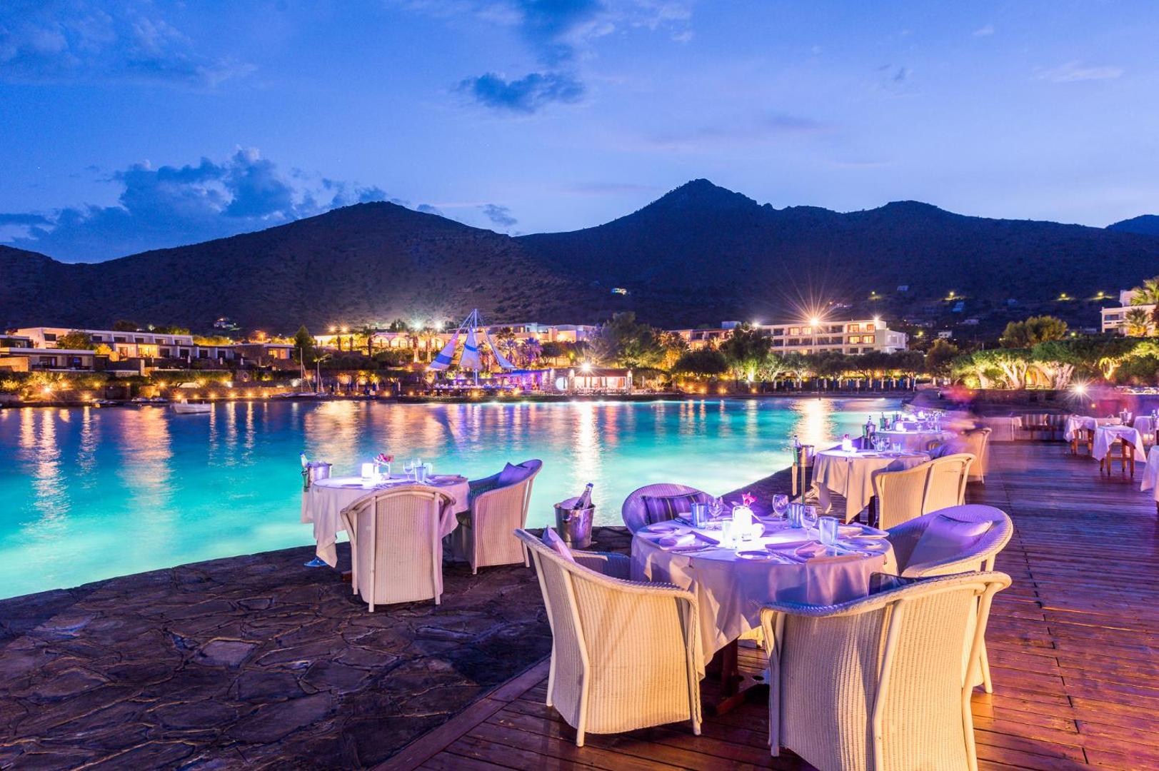 Elounda Bay Palace Hotel ēdienkartes restorāns uz terases vakarā ar skatu uz izgaismotu līci un kalniem