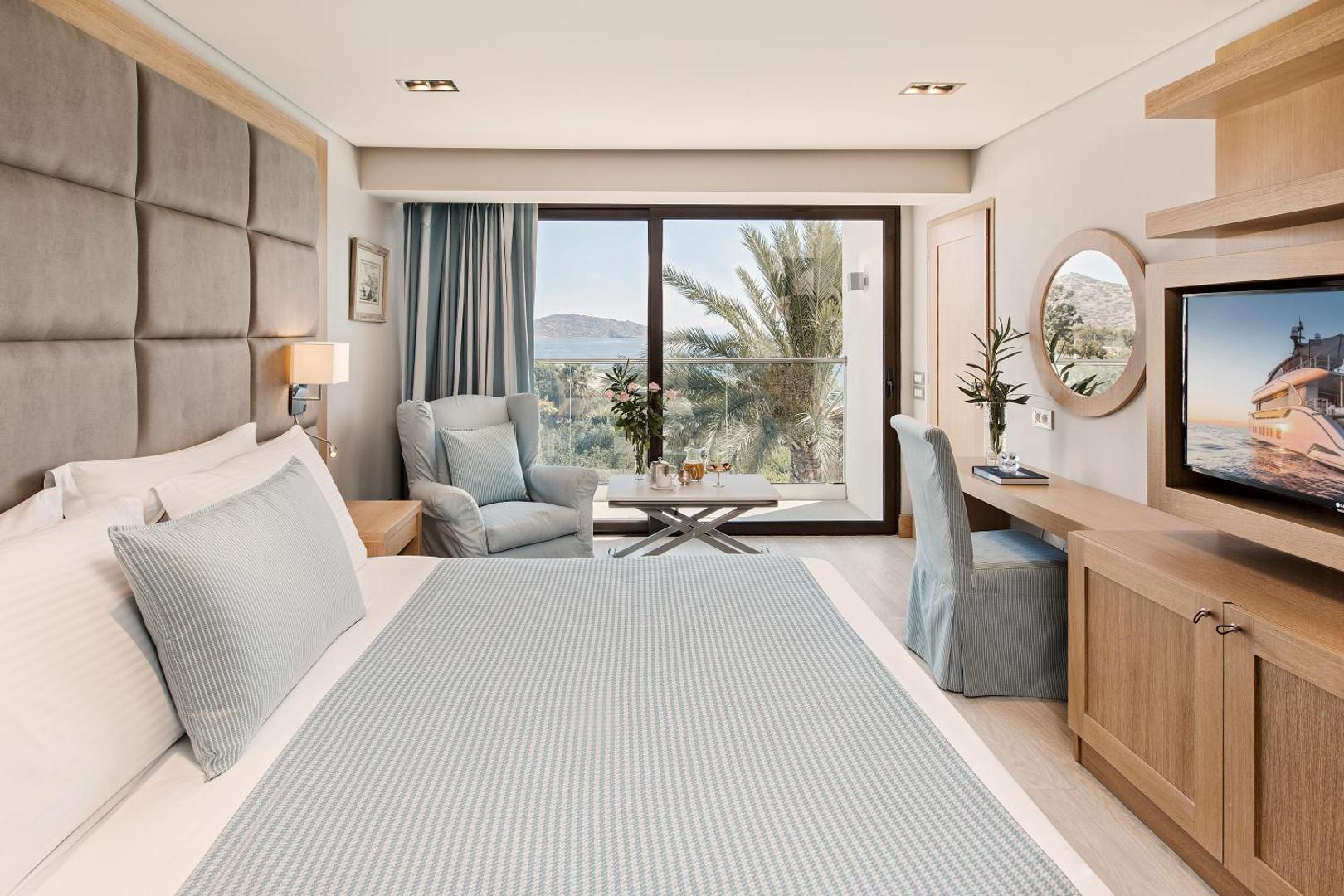 Elounda Bay Palace Hotel elegants numurs ar panorāmas logu uz palmu dārzu un jūru — mīkstais atpūtas krēsls un darba zona