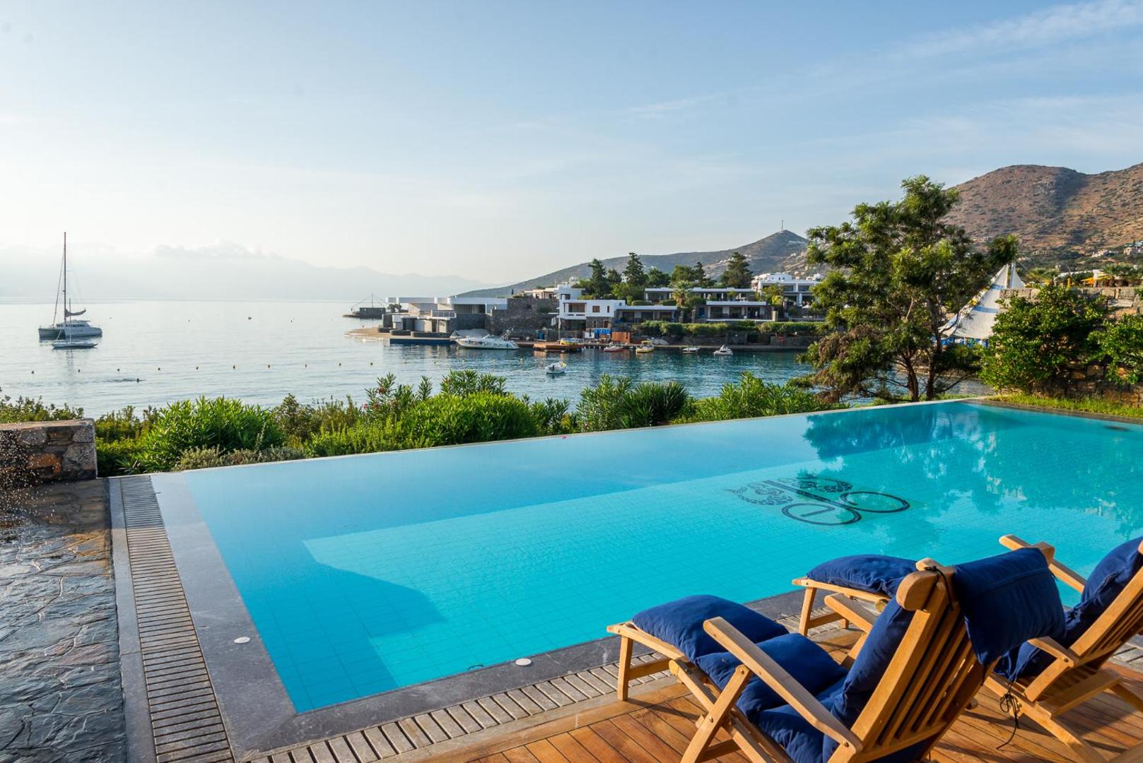 Elounda Bay Palace Hotel baseins ar pārplūdes malu un skatu uz Mirabello līci Krētā — koka terase ar atpūtas krēsliem