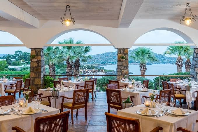 Elounda Bay Palace Hotel iekštelpu restorāns ar arkveida logiem un skatu uz jūru un palmām Krētā