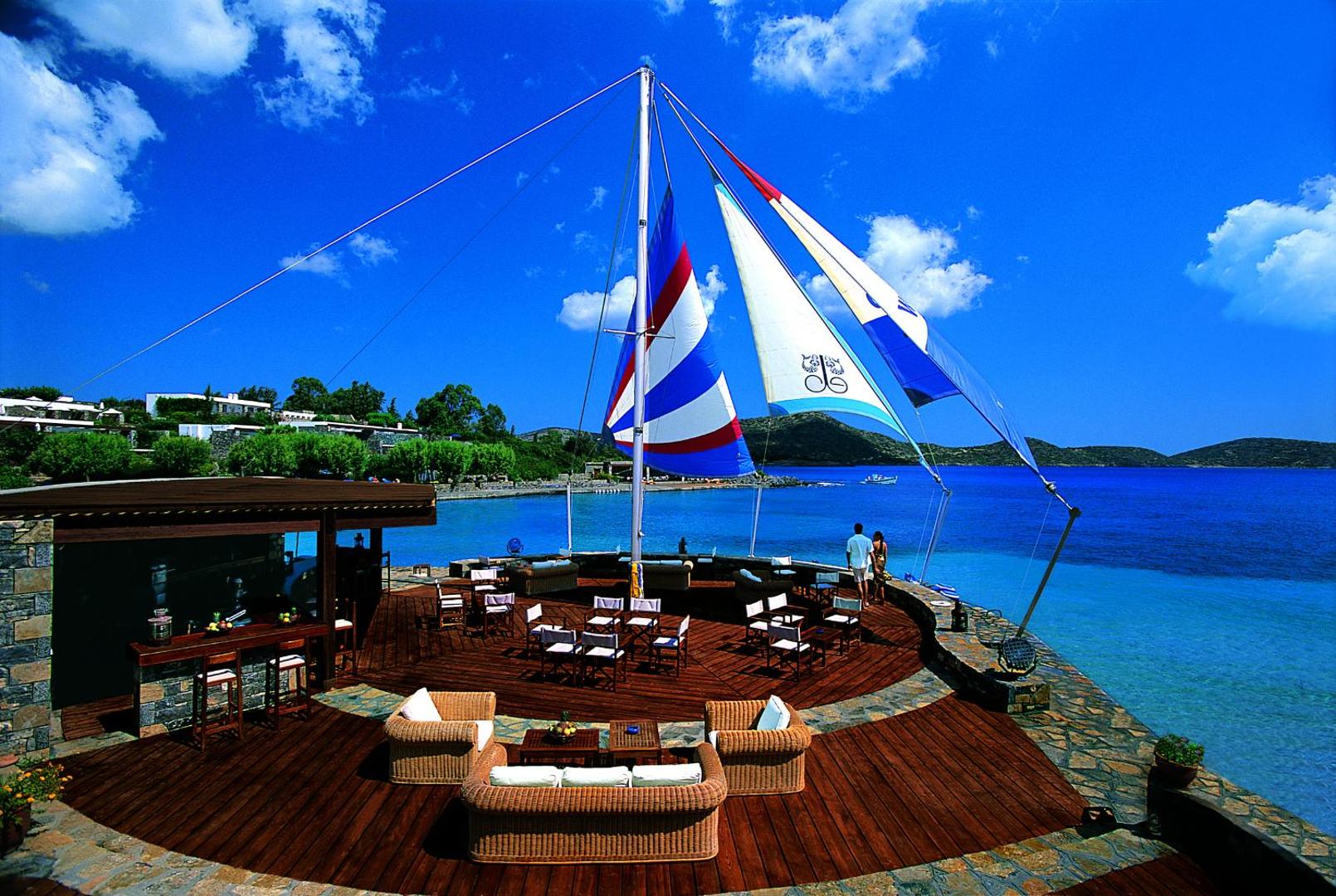 Elounda Bay Palace Hotel āra bārs uz koka terases pie jūras Krētā ar buru dizaina elementiem un atpūtas mēbelēm