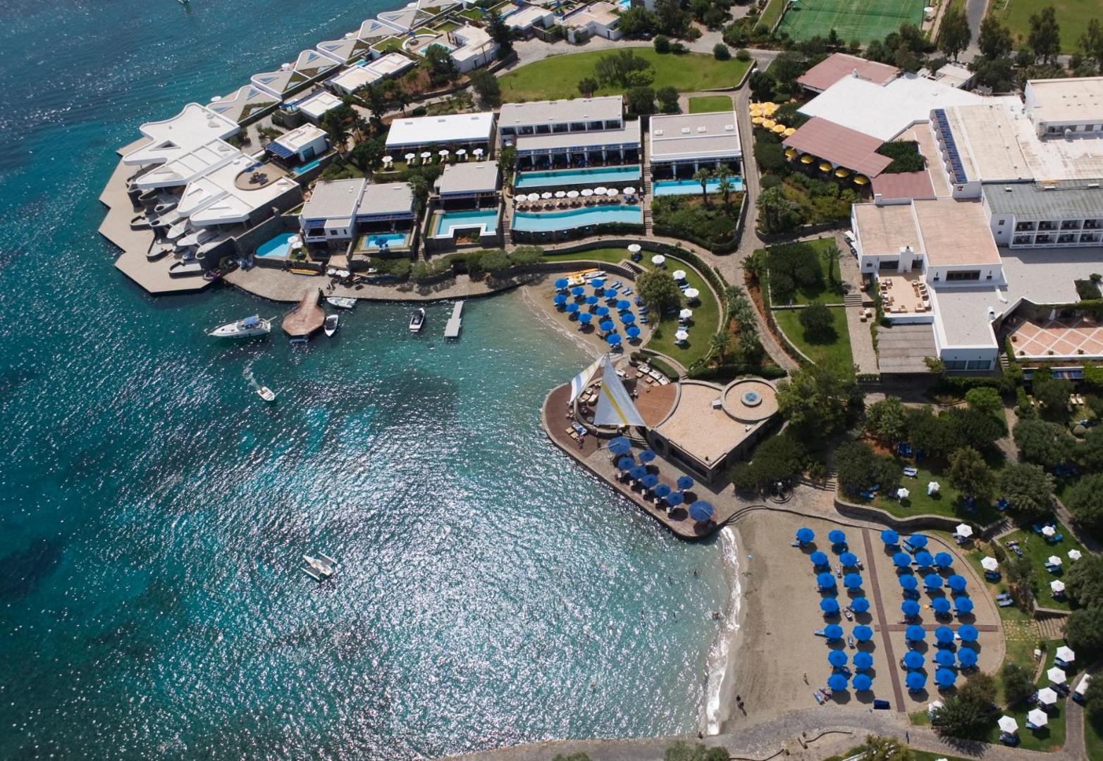 Elounda Bay Palace Hotel panorāmas skats uz jahtu piestātni, pludmali un viesnīcas ēkām saulainā dienā Krētas piekrastē.