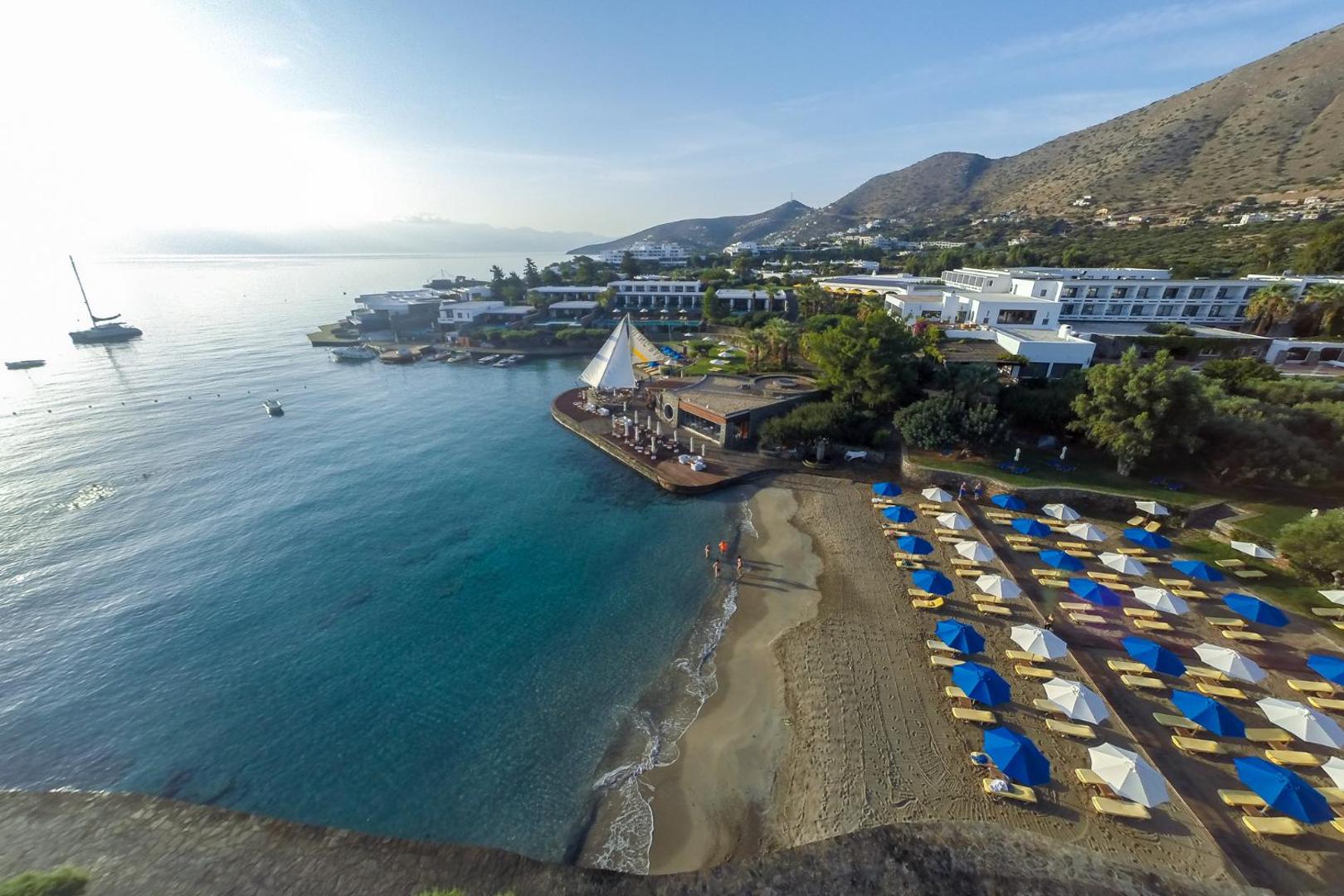 Elounda Bay Palace Hotel pludmales zona saulrietā ar kalnu fonu un rindu saulessargu pie mierīgā jūras līča.
