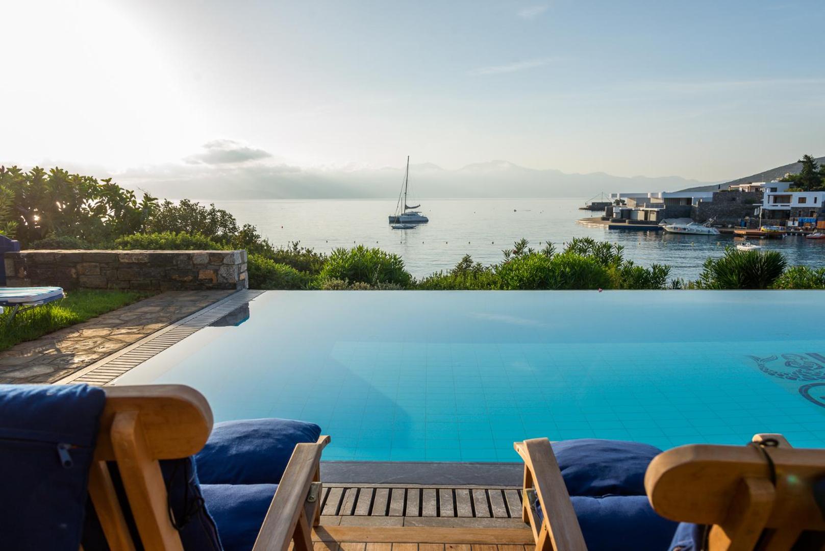 Skats no Elounda Bay Palace Hotel terases uz jahtu līcī saulrieta laikā — priekšplānā baseins ar bezgalības malu
