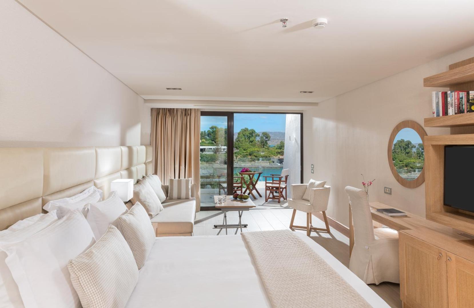 Elounda Bay Palace Hotel moderns numurs ar terasi un skatu uz baseinu un jūru — gaišs interjers ar koka elementiem
