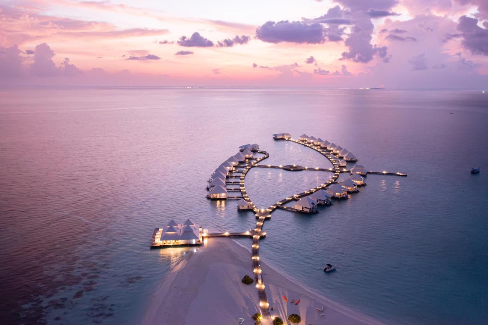 Diamonds Athuruga Maldives ūdens bungalo saulrietā ar izgaismotām koka laipām virs Indijas okeāna rozā debesu fonā.