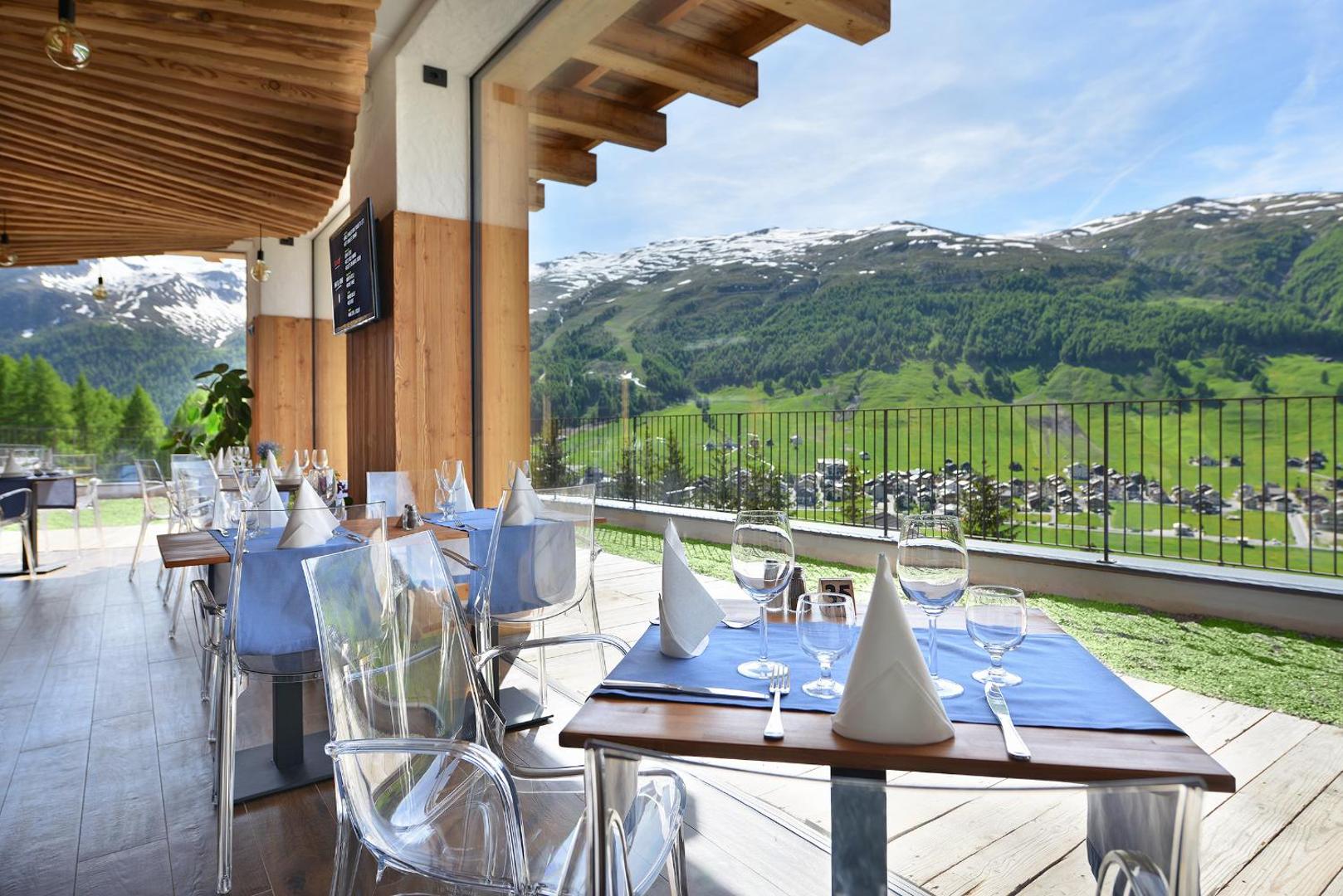 Alpen Resort Bivio restorāna āra terase ar klātiem galdiem un panorāmas skatu uz Alpu kalniem un ciematu lejā
