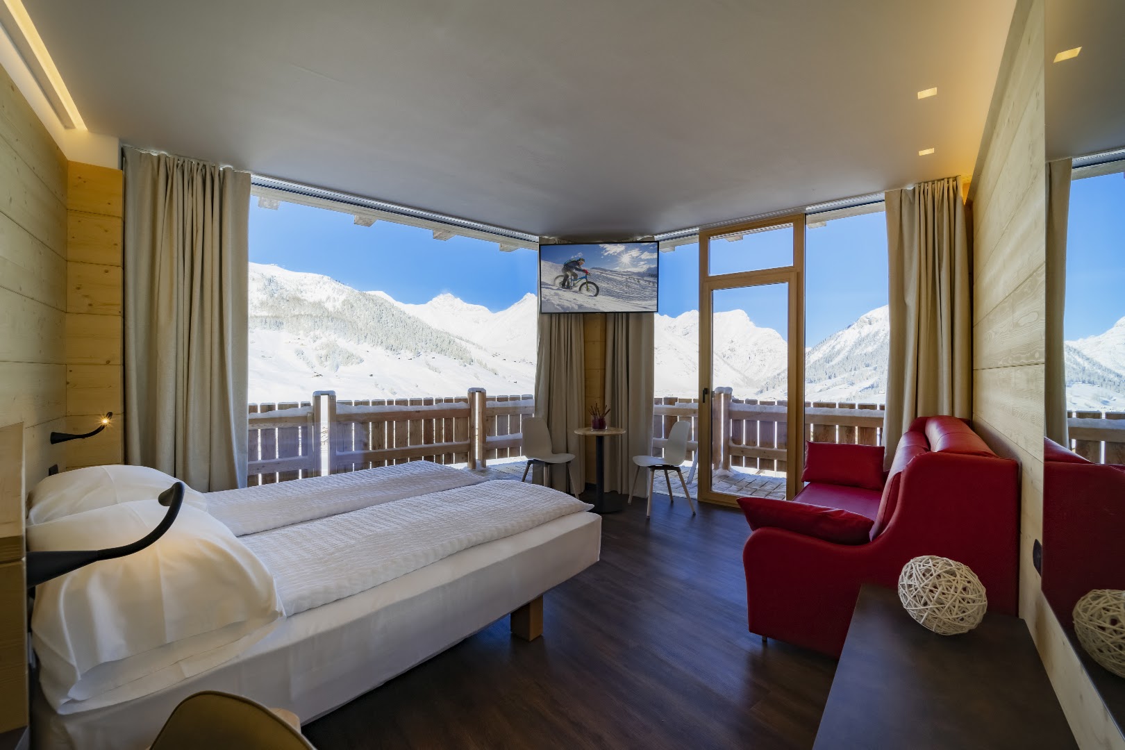 Alpen Resort Bivio gaišs numurs ar panorāmas logiem uz sniegotajām Alpu virsotnēm, koka apdari un sarkanu dīvānu atpūtai