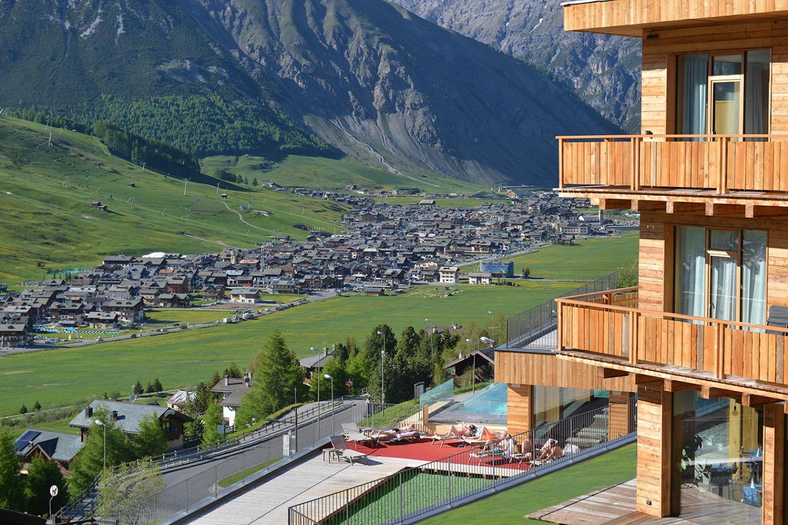 Alpen Resort Bivio skats no terases uz Livinjo ieleju vasarā — koka balkoni, sauļošanās zona un Alpu kalnu panorāma Itālijā