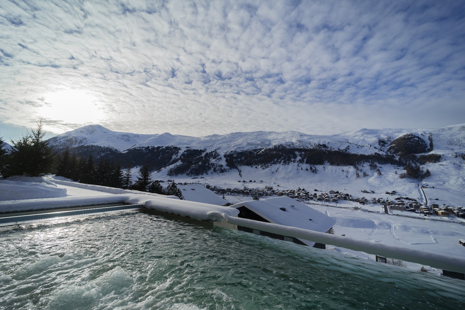 Skats no Alpen Resort Bivio džakuzi zonas uz sniegoto Livinjo ciematu un kalnu nogāzēm saulainā ziemas dienā