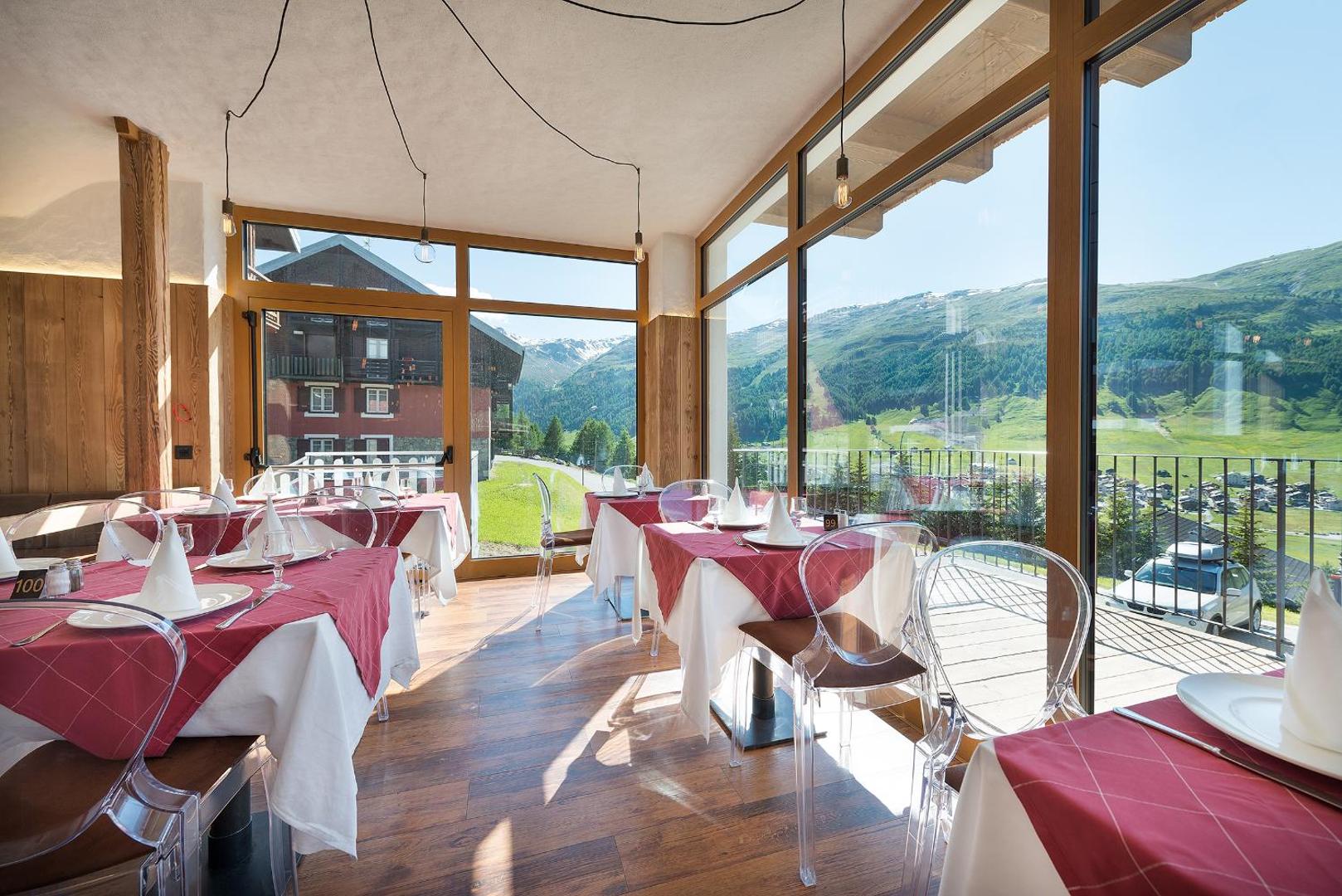 Alpen Resort Bivio gaišs restorāns ar lieliem panorāmas logiem, klātiem galdiem un skatu uz kalnu ainavu saulainā dienā