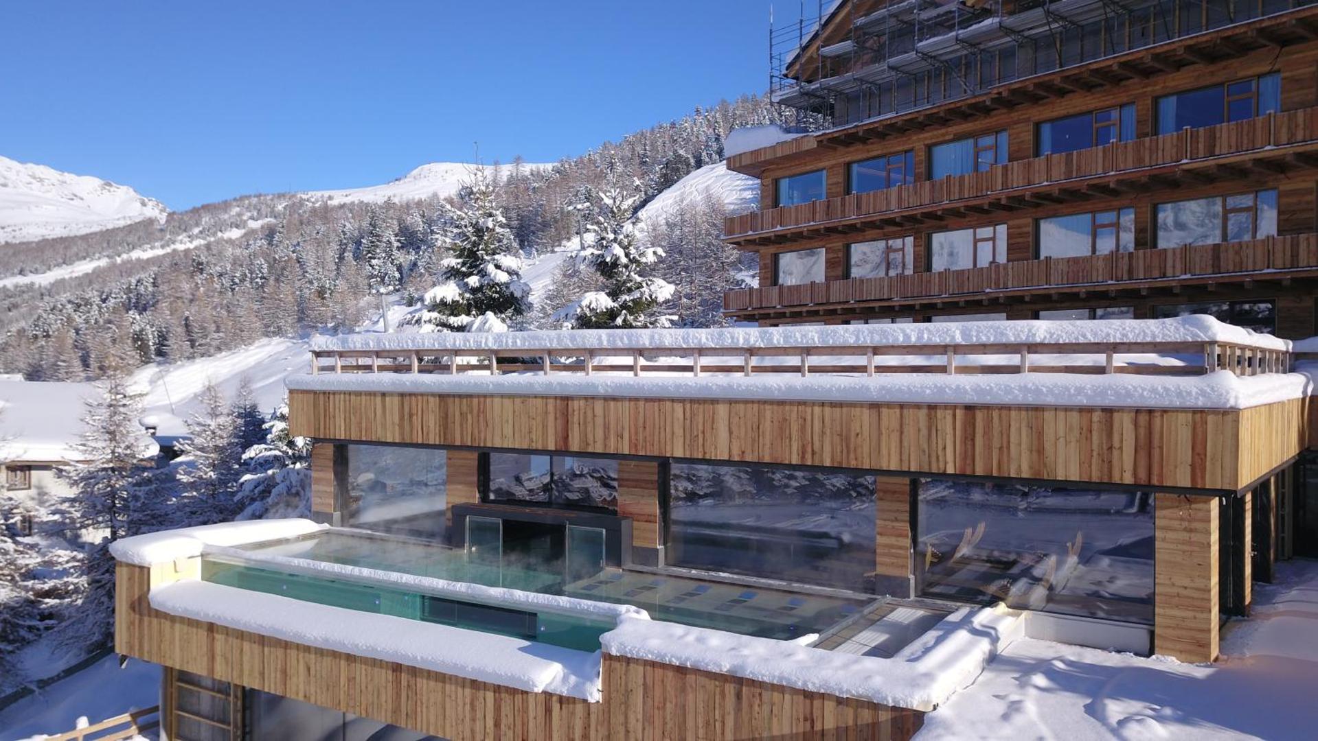 Alpen Resort Bivio apsildāmais āra baseins ziemā ar sniegotām nogāzēm fonā — moderna koka arhitektūra un Alpu ainava