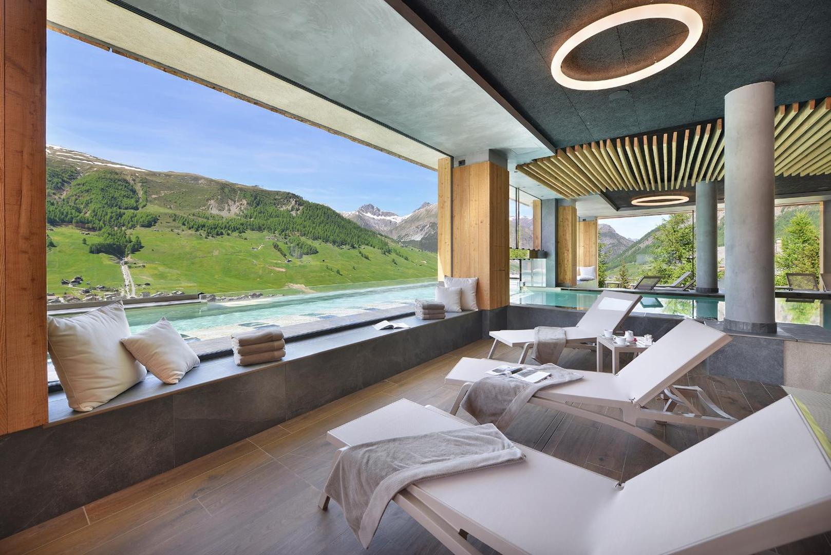 Alpen Resort Bivio spa atpūtas zona ar zviļņiem pie liela loga, no kura paveras skats uz zaļām kalnu nogāzēm