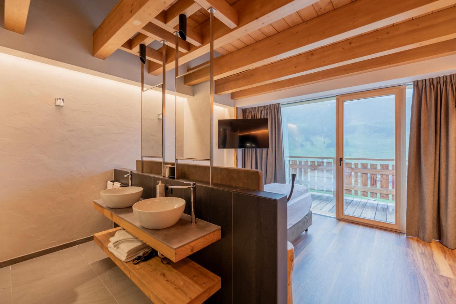 Alpen Resort Bivio numura vannasistabas zona ar dubulto izlietni, koka apdari un skatu uz zaļojošām kalnu pļavām vasaras sezonā