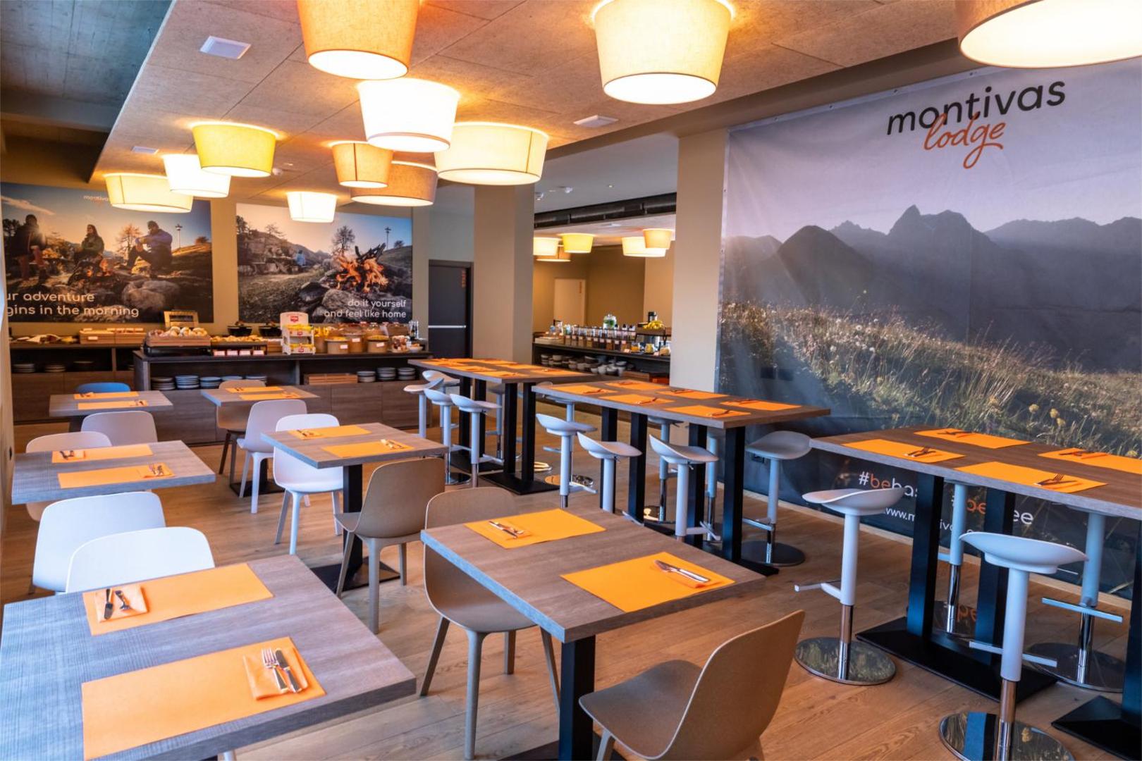 Montivas Lodge brokastu zāle ar modernām mēbelēm, oranžiem akcentiem un bufetes zonu, radot mājīgu vidi rīta maltītei.
