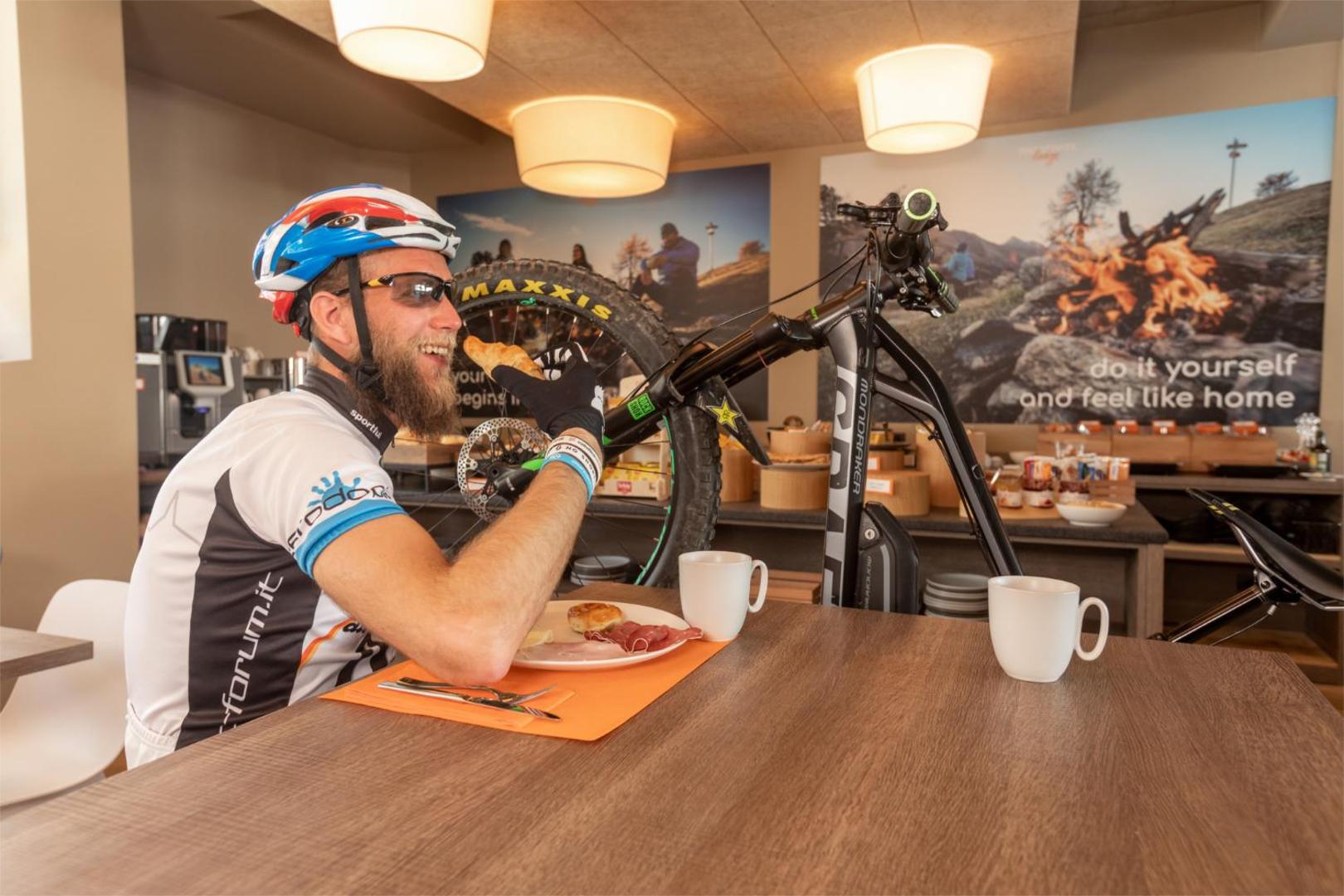 Vīrietis ar velosipēda ķiveri bauda brokastis Montivas Lodge restorānā, fonā redzams kalnu divritenis un bufetes zona Livinjo kūrortā