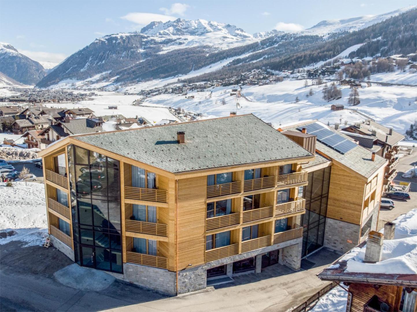 Montivas Lodge viesnīcas ēka ziemā Livinjo kūrortā Itālijā — skats no augšas uz koka fasādi un sniegotajām Alpu virsotnēm
