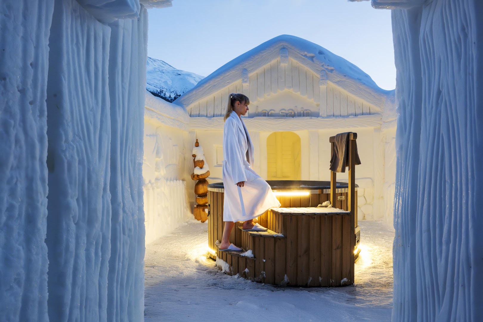 Sieviete baltā halātā pie koka kubla sniega mājiņā Lac Salin Spa & Mountain Resort teritorijā Livinjo kalnu ielenkumā