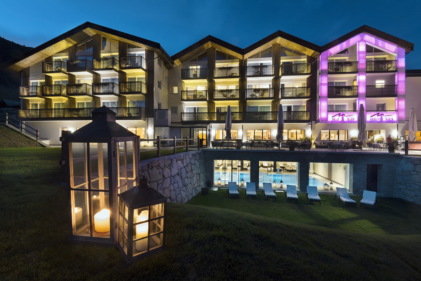 Lac Salin Spa & Mountain Resort fasāde vakarā Liviņjo, Itālijā — izgaismoti balkoni, āra terase un skats uz iekštelpu baseinu