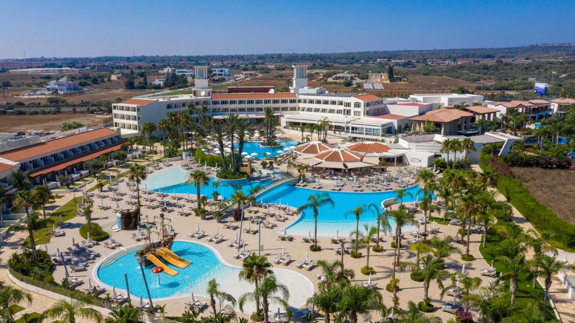 Kanika Olympic Lagoon Resort panorāmas skats uz plašo baseinu kompleksu un viesnīcas korpusiem Ayia Napa, Kiprā — saulaina...