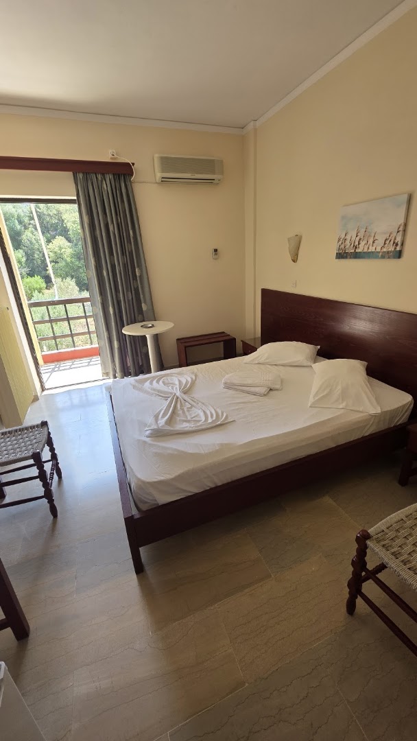 Brati Arkoudi Hotel divvietīgs numurs ar tumša koka gultu, balkonu un skatu uz dārzu Arkoudi kūrortā, Grieķijā.