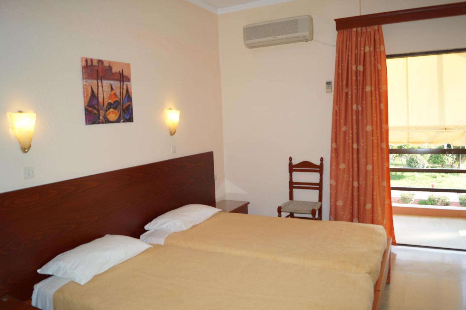 Brati Arkoudi Hotel gaišs numurs ar divām gultām, koka galvgali un izeju uz balkonu Arkoudi ciemata piekrastē, Grieķijā.