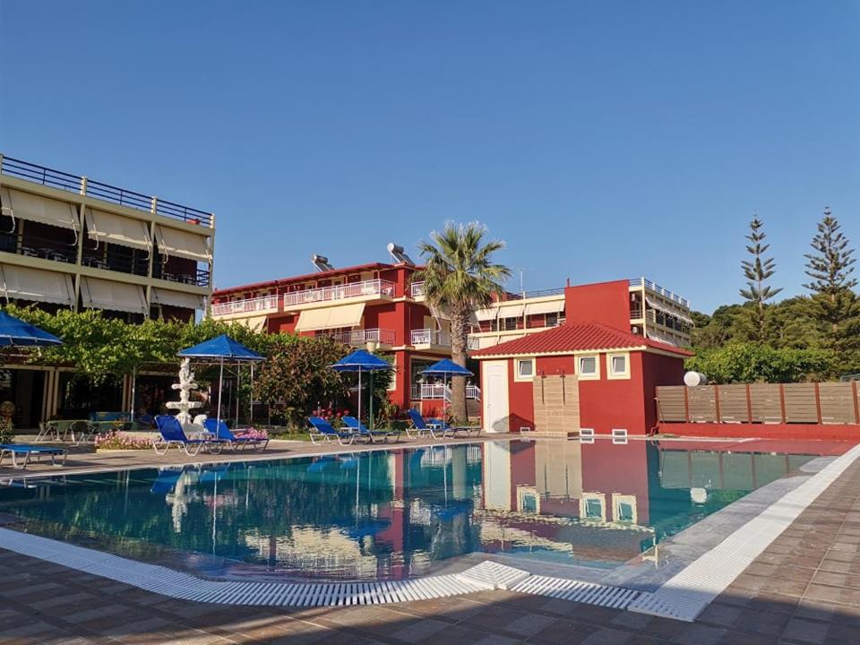 Brati Arkoudi Hotel āra baseins ar ziliem saulessargiem un guļamkrēsliem pie sarkanām viesnīcas ēkām Peloponēsas reģionā, Grieķijā.
