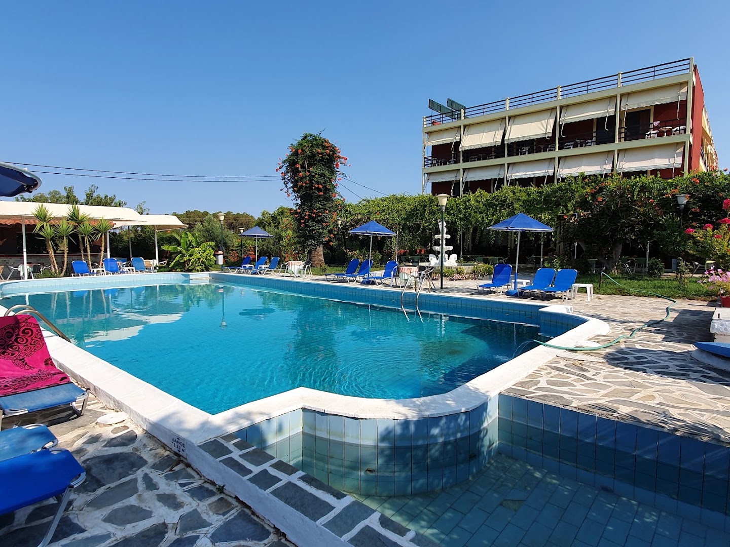 Brati Arkoudi Hotel baseina mala ar akmens flīžu klājumu, guļamkrēsliem un skatu uz viesnīcas korpusu zem skaidrām debesīm.