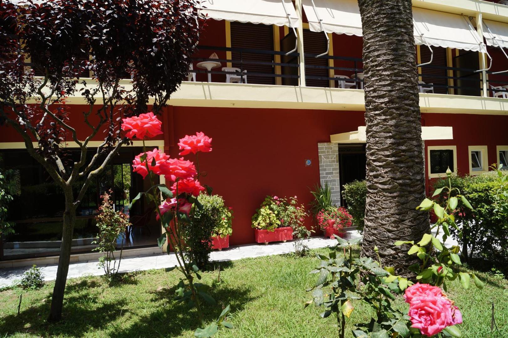 Ziedošas rozes un palmas stumbrs Brati Arkoudi Hotel dārzā pie numuru terasēm — mājīga atpūtas zona