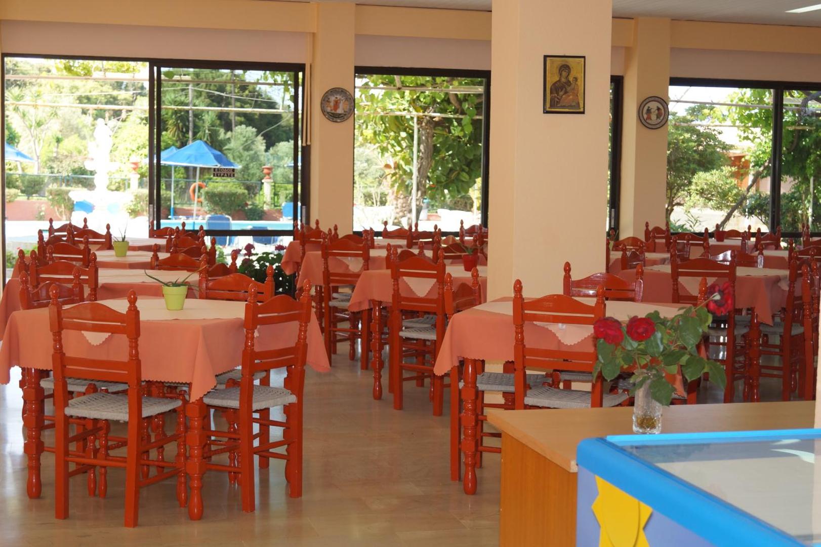 Brati Arkoudi Hotel restorāna iekštelpas ar koka galdiem un krēsliem Grieķijā — skats caur logiem uz dārzu un baseina zonu